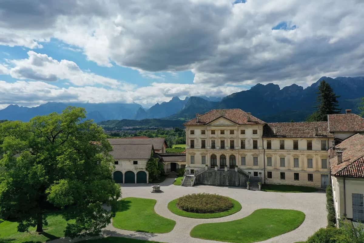 A historic noble villa with formal gardens, the Dolomites beyond, under a dramatic sky