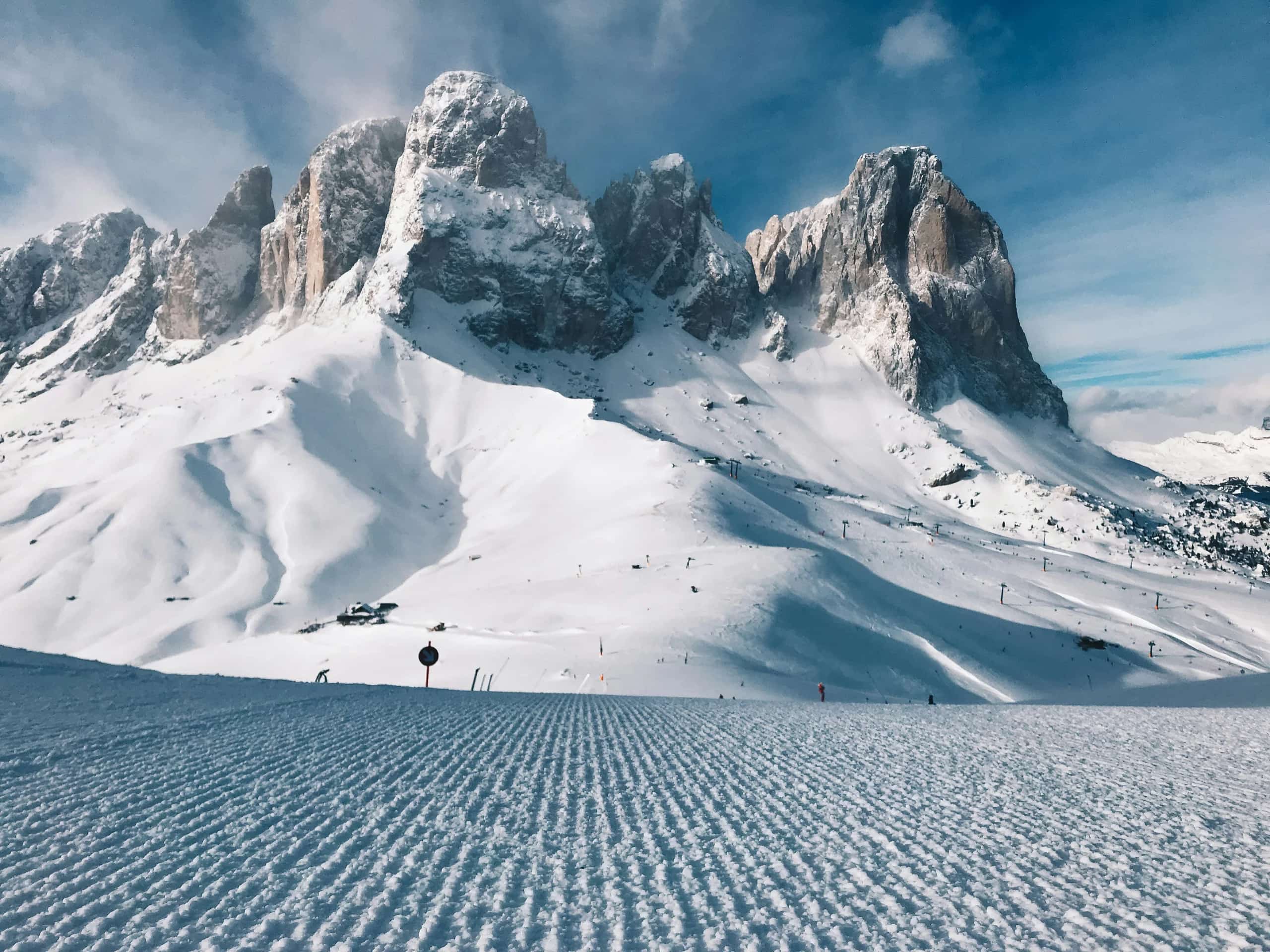 A freshly groomed ski piste beneath snow-covered Dolomite peaks under a blue sky